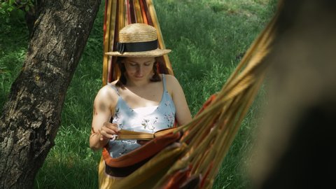 Young lady in hat with book in hammock. Charming happy woman resting in hammock and reading book at green garden. Brunette female lying in hammock with book in hands, relaxing and enjoying vacation