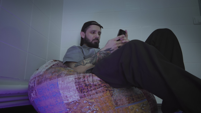 Young bearded man wearing cap texting on his smartphone while sitting on frameless chair
