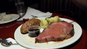 This close up video shows anonymous hands cutting into a fresh baked potato with a juicy prime rib meal at a restaurant.