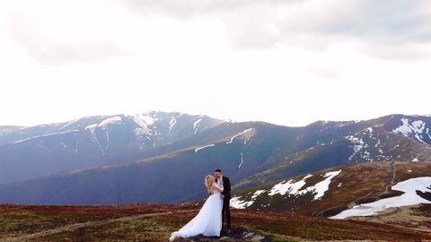 amazing aerial view, the bride approaches the groom. in a white wedding dress. standing on a background of beautiful mountains landscape. Wedding couple hugging and kissing. top of snow covered rock.