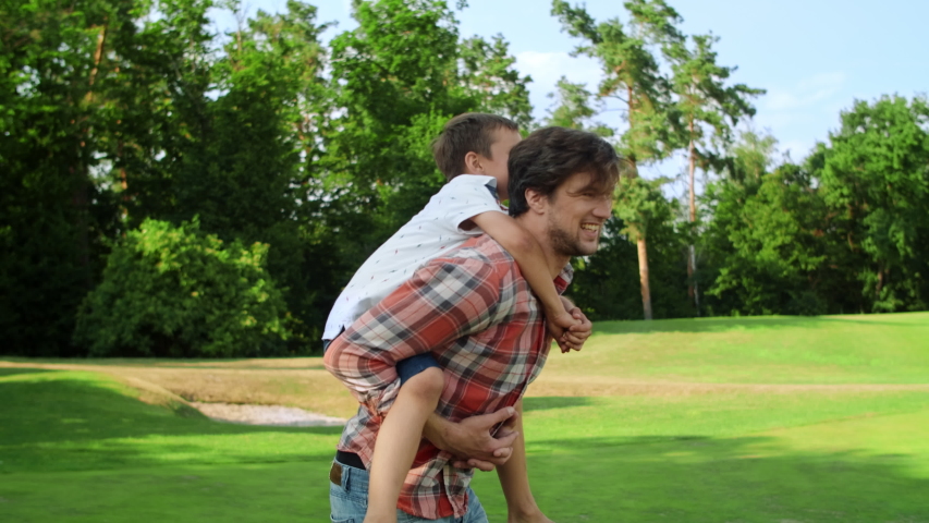 Happy father spinning around in park with son on back outside. Smiling man carrying boy on back in green park. Closeup joyful family having fun together in summer park Royalty-Free Stock Footage #1054938011