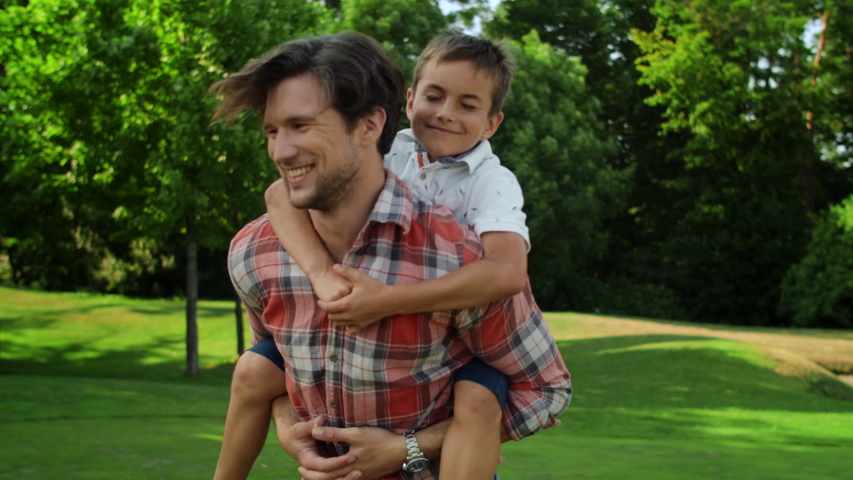 Happy father spinning around in park with son on back outside. Smiling man carrying boy on back in green park. Closeup joyful family having fun together in summer park Royalty-Free Stock Footage #1054938011
