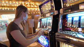 This video shows the side view of a young woman playing slot machines in an empty casino with a medical face mask, pushing the button to place her bet and clapping and celebrating when she wins big!