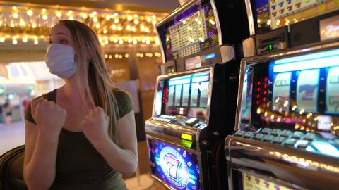 This video shows the side view of a young woman playing slot machines in an empty casino with a medical face mask, pushing the button to place her bet and clapping and celebrating when she wins big!