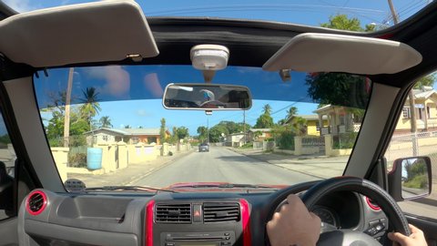 BRIDGETOWN, BARBADOS, CARIBBEAN ISLANDS, DECEMBER 2019: POV: Unrecognizable man drives you in a red Suzuki Jimny through the tropical suburbs. Male tourist explores Bridgetown during his vacation.