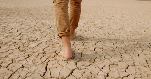 Close up shot of feet of adult man walking barefoot on bottom of dried lake or river, stepping on cracked soil ground destroyed by erosions - ecological issues concept 4k footage