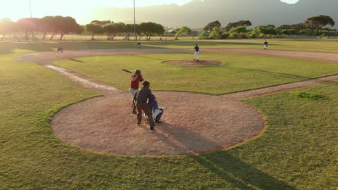 Members of a baseball team playing a game outdoors on a baseball field at sundown, the hitter hitting a pitch and making a run, seen from behind the catcher, shot with drone