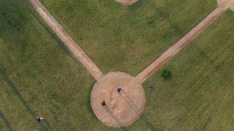 Overhead view of a baseball team playing a game outdoors on a baseball field at sundown on a sunny day, the hitter making a run and arriving back at home base as fielder catches ball, shot with drone