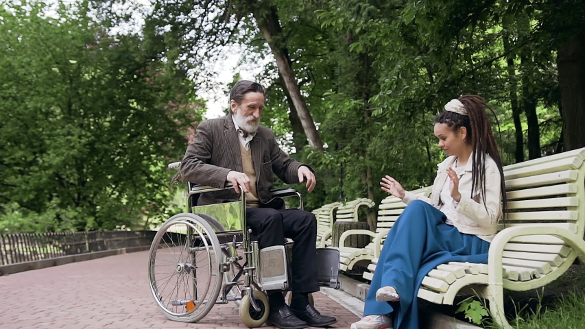 Splendid surprised stylish 25-aged girl with long dreadlocks helping to get up her respected confident senior grey-bearded grandfather from wheelchair during joint walking in urban green park