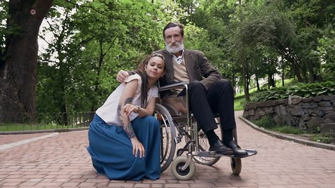 Family concept where high-spirited adorable friendly loving granddaughter with tatoo on arm and long dreadlocks sitting near her esteemed smiling senior grandfather in wheelchair in green park and