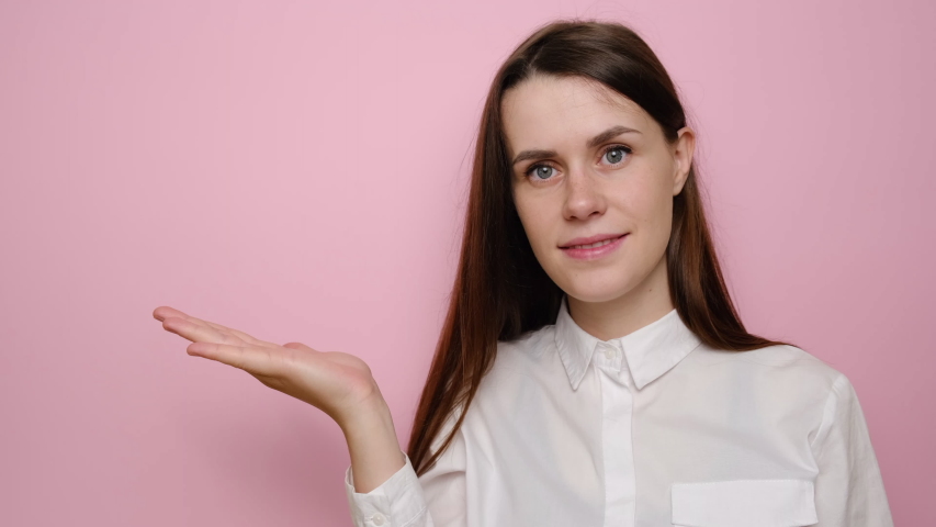 Smiling pretty young woman looks pleasant to her palm hand, showing something in her hand, dressed in white shirt, isolated on pink studio background with copy space. People sincere emotions concept