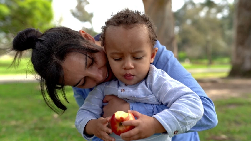 Little boy eating red apple, chewing and sitting on mom knees. Mixed-race young mother talking with him in city park. Childhood and weekend concept Royalty-Free Stock Footage #1054954412