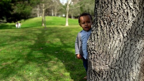 Happy African American little boy running around tree. Cute toddler playing hide-and-seek in city park at summer. Childhood and weekend concept