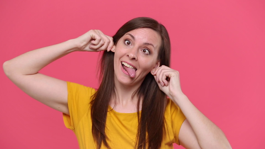 Fun young woman in yellow t-shirt posing isolated on pink background studio. People lifestyle concept. Looking camera fooling around pointing index fingers on blowing cheeks monkey ears showing tongue Royalty-Free Stock Footage #1054954847