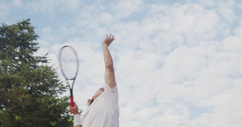 A Caucasian man wearing tennis whites spending time on a court, playing tennis on a sunny day, holding tennis racket, hittig a tennis ball, in slow motion.
