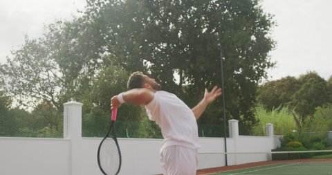 A mixed race man wearing tennis whites spending time on a court, playing tennis on a sunny day, holding tennis racket, hittig a tennis ball, in slow motion.