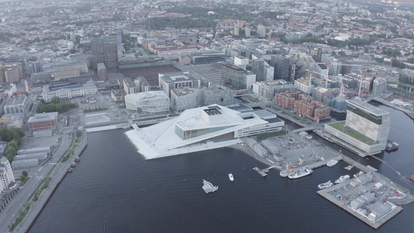 Oslo, Norway. The Oslo Opera House dominates the waterfront with its angular white marble and granite form rising directly from the fjord, its sloping roof creating a continuous public plaza that merges architecture with landscape. The surrounding Bjorvika district frames the scene with contemporary high-rise buildings and active harbor infrastructure, while boats move through the dark water in the foreground. Warm sunset backlight casts long golden rays across the cityscape, illuminating the white surfaces of the opera house and contrasting with the deep blue-gray tones of the water and urban fabric. The aerial perspective reveals the full geometric footprint of the landmark and its relationship to the revitalized waterfront, capturing a moment of transition between day and evening in the Norwegian capital. The building