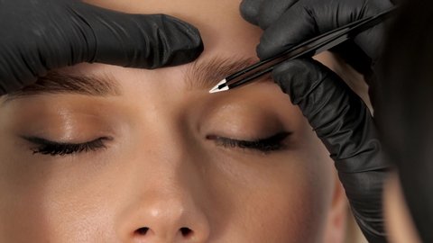 Stylist's hands in black gloves plucking eyebrows with tweezers. Beautiful attractive female face of a blonde well-groomed woman or lady. Styling and lamination of eyebrows.