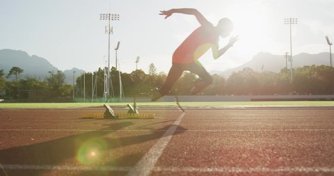 Fit, disabled mixed race male athlete with prosthetic legs on a running track at an outdoor sports stadium starting a race from starting blocks, in slow motion backlit with lens flare