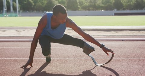 Fit, disabled mixed race male athlete with prosthetic legs on a running track at an outdoor sports stadium, stretching on the track in the sun, in slow motion