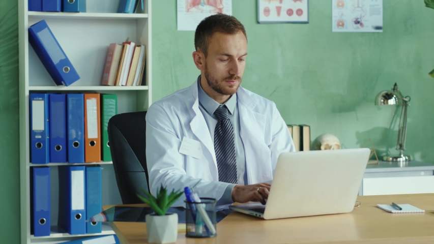 Medium shot european doctor in the office consulting people via internet using laptop. Indoor portrait smiling confident family therapist in white gown at work. Royalty-Free Stock Footage #1054967573