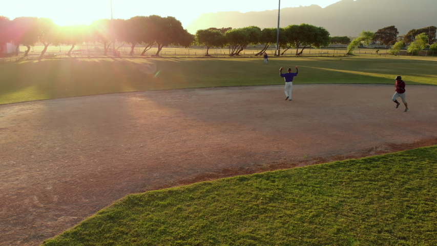 Members of a baseball team playing a game outdoors on a baseball field at sundown on a sunny day, the hitter making a run and arriving back at home base, shot with drone Royalty-Free Stock Footage #1054970195