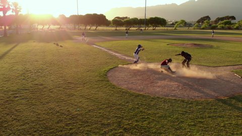 Members of a baseball team playing a game outdoors on a baseball field at sundown on a sunny day, the hitter making a run and arriving back at home base, shot with drone