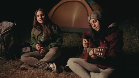 dolly tracking shot.
Group of young people having fun sitting near bonfire outdoors at night playing guitar, singing songs and dancing, drinking coffee or tea. Campfire, tent, young girls in nature