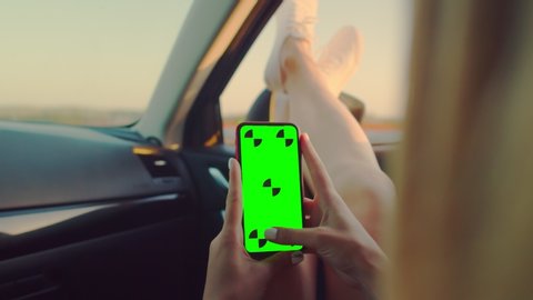 Women's hands hold a phone with a green screen with their hands on the steering wheel of the car. The girl driver is sitting at the wheel holding a phone with a chromakey in her hands