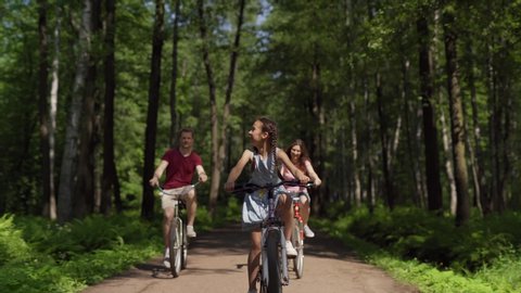 Happy family of father, mother and little daughter looking around at nature while riding bicycles together in green forest. Playful girl enjoying cycling faster than parents