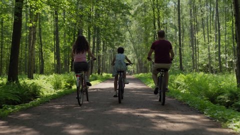Back view follow shot of happy family of father, mother and little daughter riding bicycles together down trail in green forest