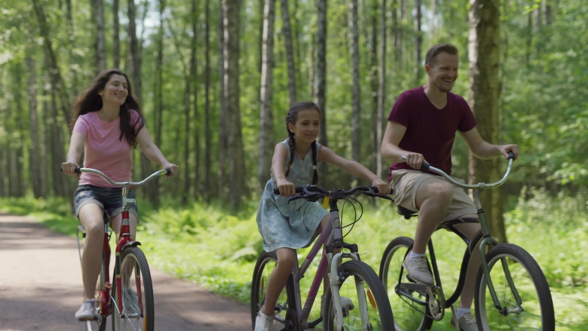 Tracking shot of happy family of mother, father and cute daughter enjoying cycling together down trail in forest | Shutterstock HD Video #1054977758