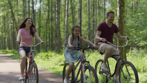Tracking shot of happy family of mother, father and cute daughter enjoying cycling together down trail in forest