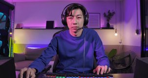 Excited asian male gamer in headset talks to other players while playing and winning in online video game on his computer in colorful neon lights room at home. eSport Cyber Games. Monitor screen view.