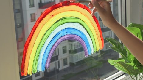 Girl painting rainbow on window during quarantine at home.