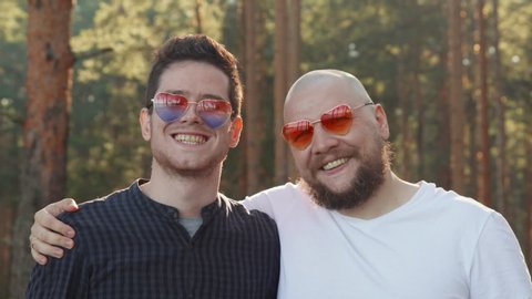 Portrait of two affectionate gay men in love wearing sunglasses of heart shape smiling to camera. Homosexual couple dating in nature. LGBTQI, Pride Event, LGBT Pride Month, Gay Pride Symbol
