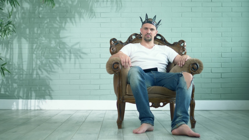 barefoot man in a t-shirt and jeans in a leather crown sits in an armchair.