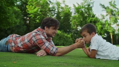 Closeup father and son lying on green grass in field. Smiling man and boy competing in arm wrestling outdoors. Positive parent and kid playing armwrestling in park. Family having fun outside