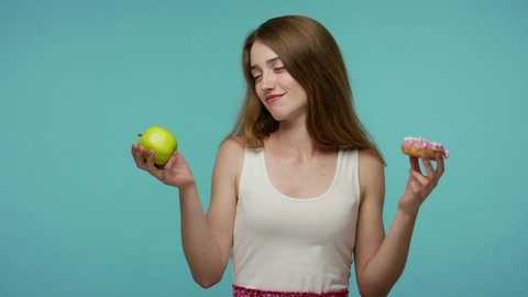 Slim beautiful girl smelling donut and biting apple, making choice between delicious fruit vs sweet sugary doughnut, vitamin nutrition concept, healthy diet. studio shot isolated on blue background