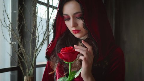 Beautiful young sexy charming woman, red velvet vintage hood stands near window gothic dark room. Backdrop branches. Girl gently touches rose petals flower. Face closeup, bright evening holiday makeup