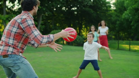 Happy family playing with ball in summer park. Positive parents throwing ball in air. Smiling children trying to catch ball in green field. Cheerful family having fun together outdoors