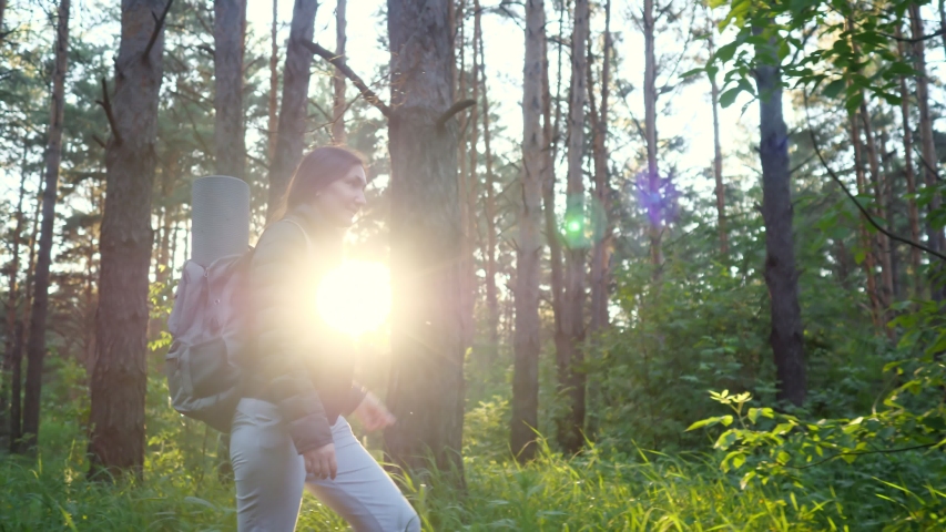 woman with a backpack walks through the coniferous forest against the backdrop of the setting sun.