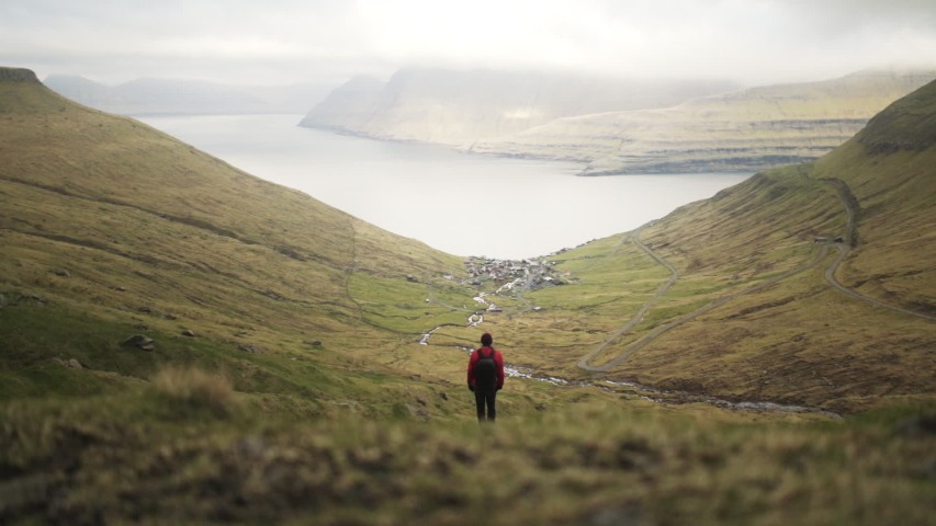 Wide Pedestal Shot Of Hiker Standing On Slope Looking Down Towards Village Of Funningur, Faroe Islands, Denmark