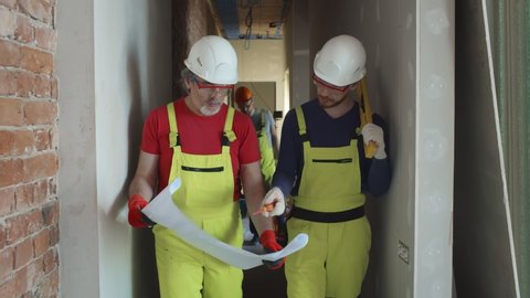 Young engineer discussing blueprint with mature building worker on construction site. Builders in protective wear study renovation blueprint in apartment