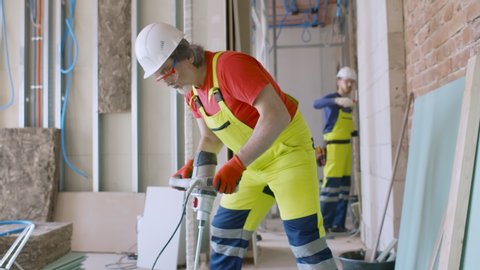 Portrait of construction worker in protective hardhat and uniform mixing tile adhesive or cement with a power drill in unfinished new apartment.