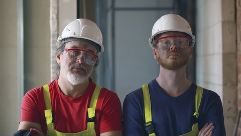 Portrait of concentrated foreman and builder standing with crossed arms on construction site. Serious professional construction workers in safety gear posing at camera