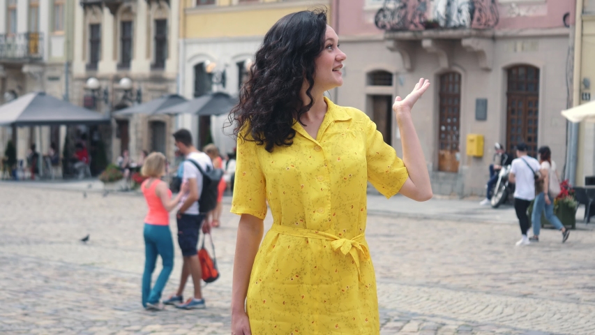 Attractive girl wearing yellow dress filming selfie video. Curly woman having video stream on holiday, waving at webcam on smartphone camera sharing travel vacation. Urban city background.