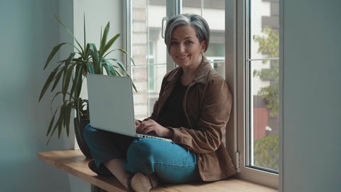 Happy mature freelancer gray haired woman working on laptop sitting leaned on the window. Woman smiling on camera in a quiet place and works sitting in a Turkish pose. Business concept. Prores 422. 