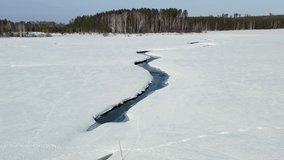Flying in winter over a swamp covered with forest. A narrow channel of dark open water cuts a winding path through a vast expanse of snow-covered ice, creating a stark contrast between the deep blue-black water and the white frozen surface. The aerial camera glides smoothly above the frozen wetland, following the serpentine waterway as it curves toward a dense treeline of mixed birch and coniferous forest on the horizon. The scene is bathed in bright, clear daylight under a pale blue sky, with the sun casting crisp shadows along the raised edges of the ice. The surrounding landscape is flat and uninterrupted, emphasizing the isolation and scale of the winter terrain. The footage captures the quiet stillness of a frozen swamp in the Ural region of Russia, where the transition between ice and water reveals the underlying river system beneath the seasonal snow. - Powered by Shutterstock - Get 15% off with code: PIKWIZARD15