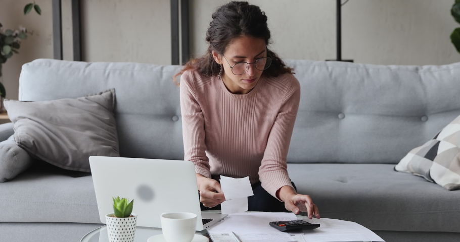 Focused serious young woman in eyewear sitting at coffee table at home, managing personal monthly budget in computer application. Concentrated lady calculating incomes outcomes utility bills.
